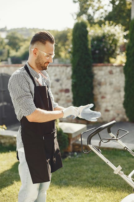 A man with glasses and a beard is seen performing garden maintenance in a small backyard. He is wearing a dark hoodie and jeans, using an electric grass trimmer to cut the grass along the edge of the